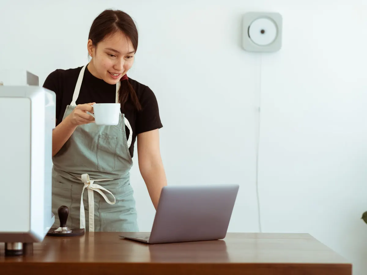 Una mujer con delantal revisa su laptop con una taza de café en la mano, en el mostrador de su café.